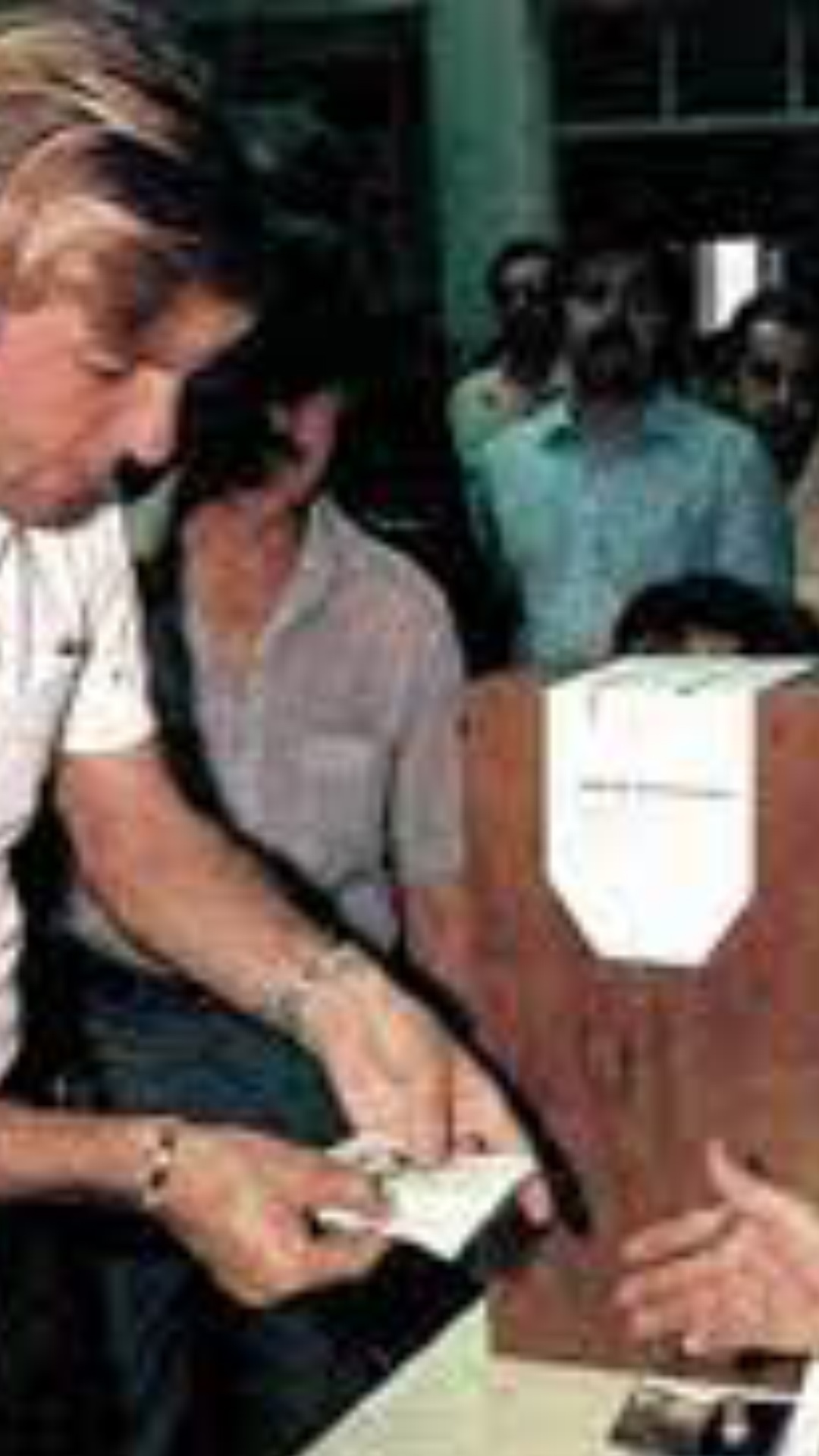 Mesa electoral del 30 de octubre de 1983. Ciudadano argentino entrega documento de identidad al presidente de mesa, quien extiende la mano. Le sigue una larga fila de votantes. 