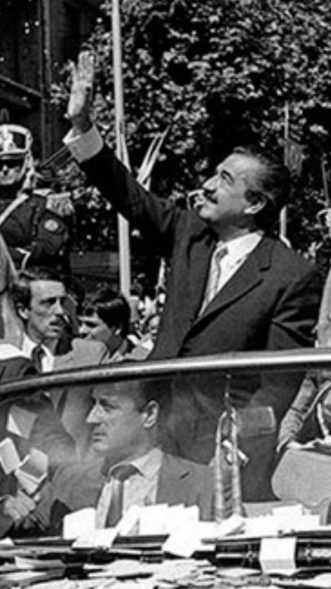 El presidente electo, Raúl Alfonsin junto a su mujer, desde un auto descapotable, saludan a una multitud. A los costados del auto es acompañado por personal de seguridad y caballeriza.