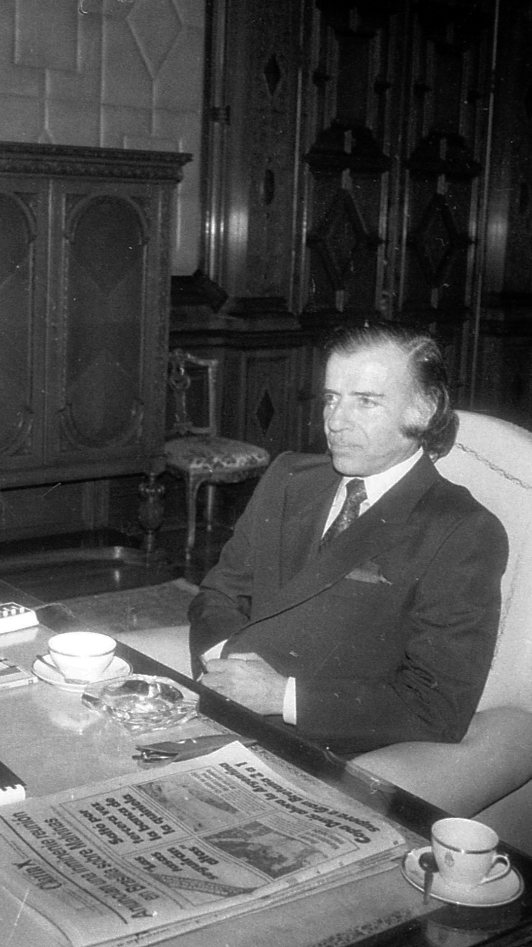 Foto muestra al presidente de ese entonces Carlos Menem junto a su ministro de economía Cavallo sentados en el despacho presidencial. EL ex presidente sentado en la punta de la mesa mirando serio al frente y Cavallo sentado a su derecha mirando a la cámara. Sobre la mesa hay una carpeta, el diario Clarín y unas tazas de café. 