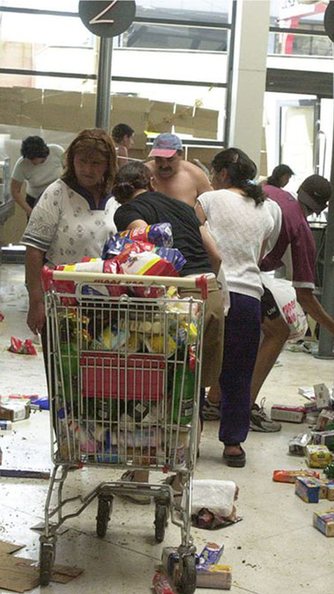 Saqueo en un supermercado en 2001. En medio de la góndola hay un carrito de supermercado lleno de productos, productos como galletitas, yerba y té desparramados sobre el piso. Tres mujeres y dos hombres se llevan cosas.