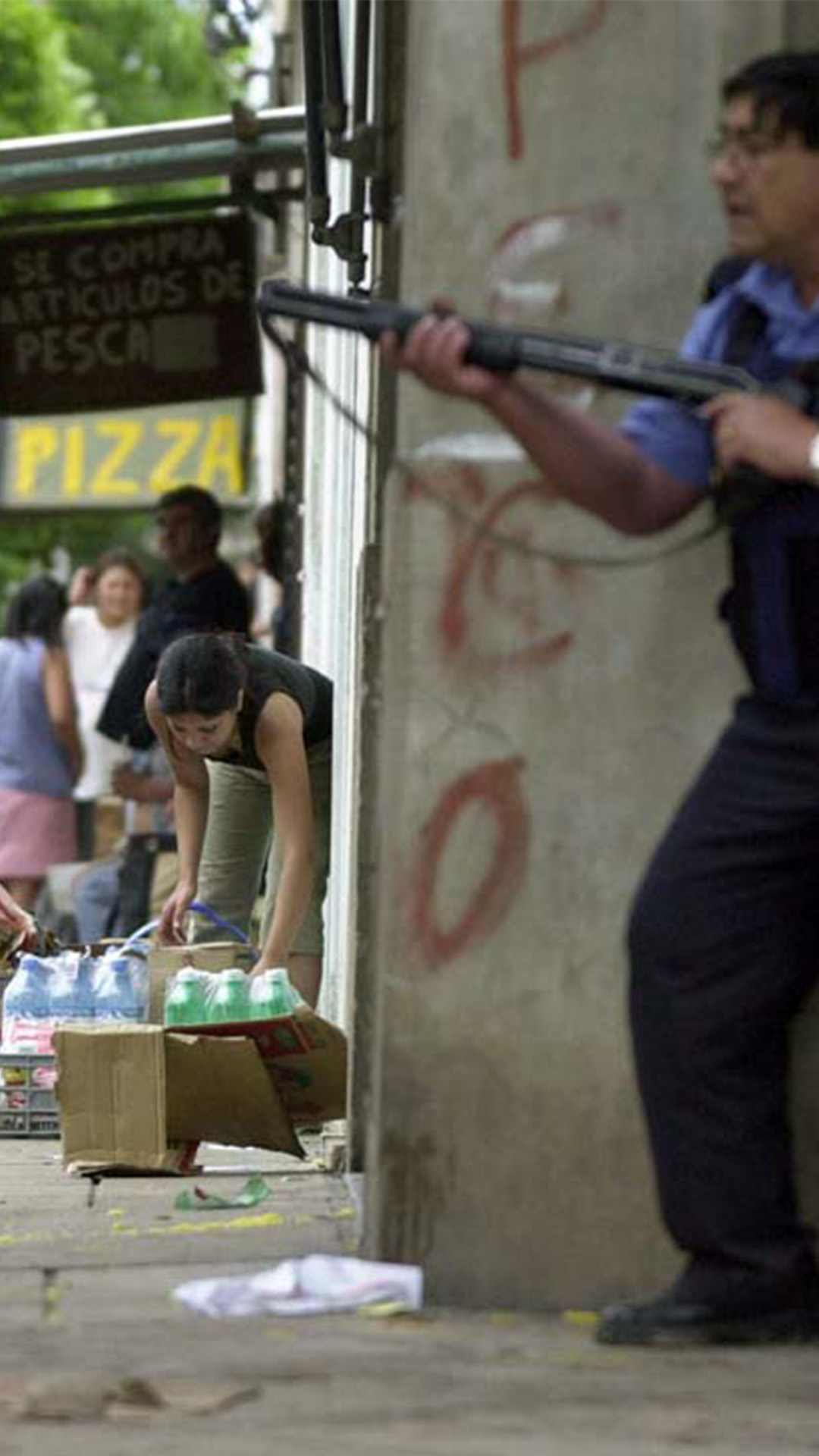 Un policía, con su arma, apostado sobre una pared apunta a la gente saqueando los supermercados. 