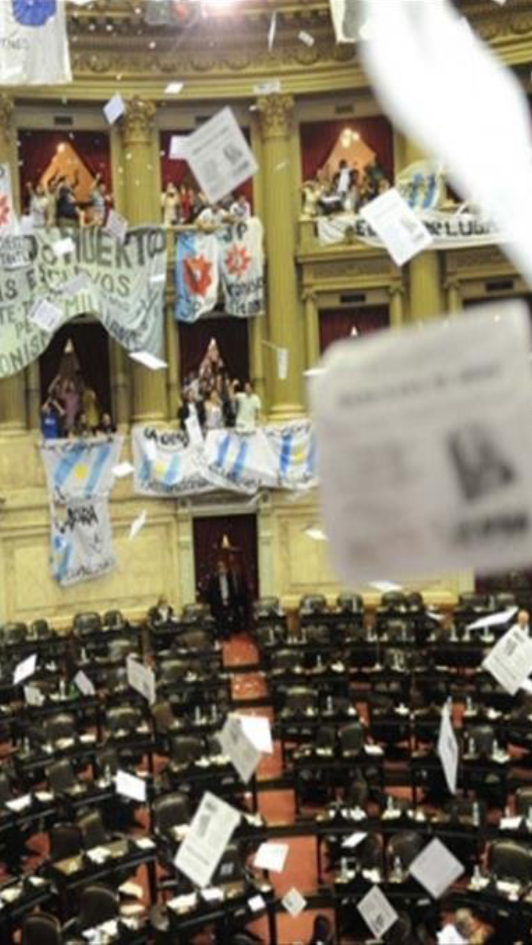 Aprobación de la Ley Voto Jóven en Diputados, el recinto está mitad vacío. En los balcones los militantes festejan, vuelan papeles de color blanco con inscripciones en negro. En los balcones se despliegan banderas.