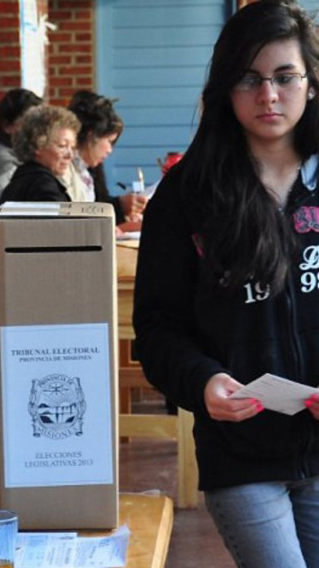 Elecciones de 2015. Una jóven se dirige a introducir su voto en la urna, en la mesa la presidenta de mesa charla con otra autoridad.