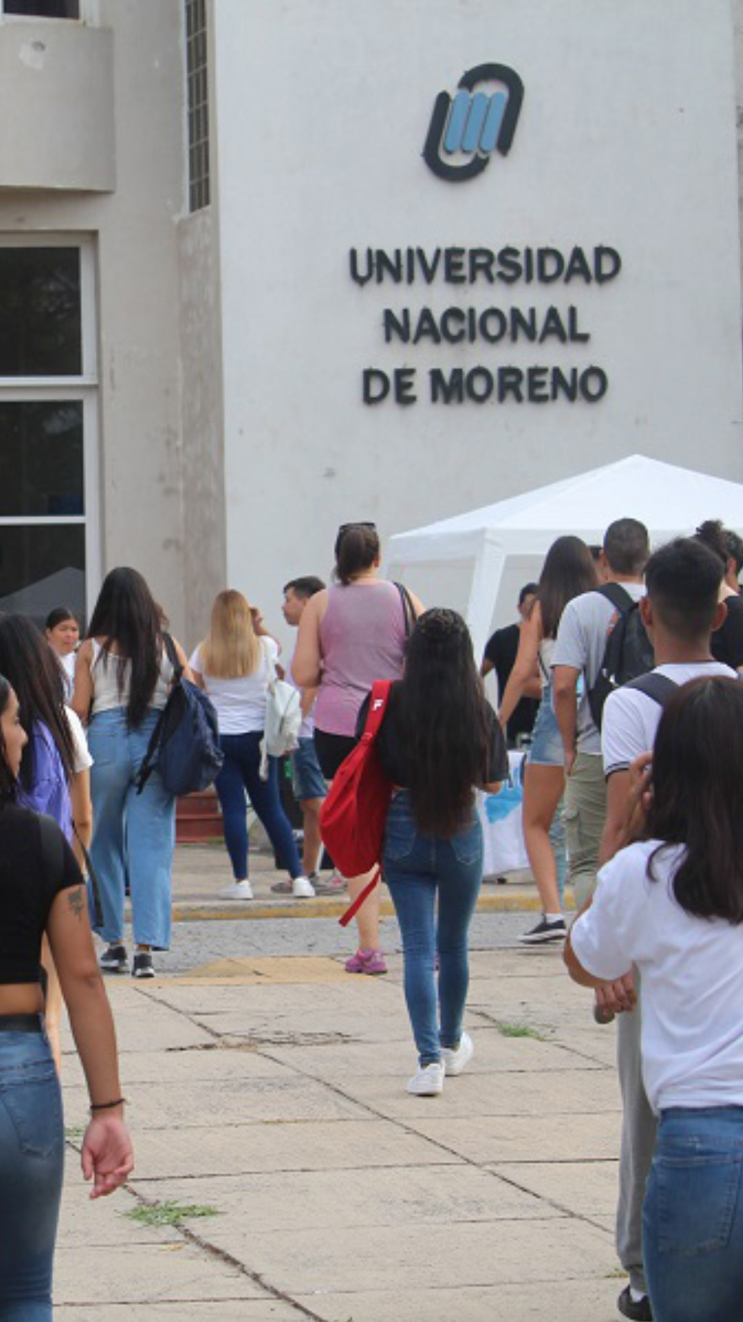 Portada de la Universidad Nacional de Moreno. La foto es exterior, de día, de frente a la entrada del edificio con varios estudiantes ingresando a la misma. Los estudiantes están de espalda a la cámara que fotografió el momento caminando hacia el edificio universitario. El establecimiento tiene el logo y el nombre de la universidad inscripto en una pared y una puerta grande, vidriada, a la cual están ingresando los alumnos.