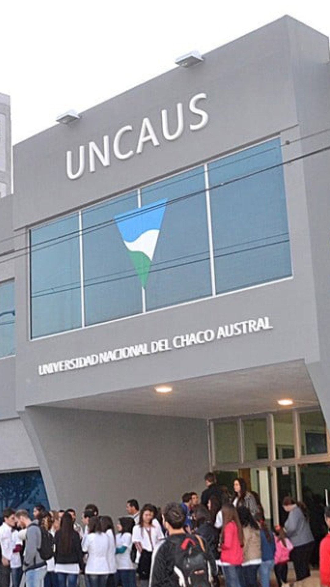Portada de la Universidad Nacional del Chaco Austral. La foto es exterior, de día y muestra el edificio desde su costado derecho con muchos estudiantes en la entrada de la institución y personas comunes caminando sobre la vereda del lugar. El establecimiento es de color gris, con muchas ventanas, con el nombre en el frente y el logo en el ventanal grande que esta sobre la entreda en el primer piso.