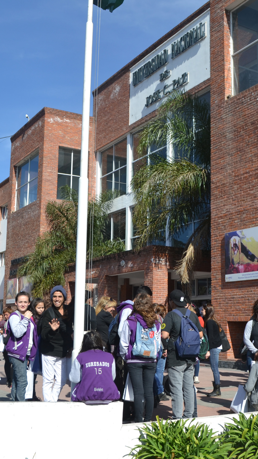 Portada Universidad Nacional de José C. Paz. La foto es exterior, de día y muestra jóvenes sentados en el playón que tiene tres mástiles con la bandera argentina y que está ubicado en la entrada de la Universidad. La toma de la foto es desde el costado derecho, es un plano general donde hay jóvenes sentados de espaldas, otros de costado y algunos mirando la cámara. De fondo aparece el edificio que tiene muchas ventanas con paredes de ladrillos a la vista y con dos cuadros de obra de arte colgados en las mismas