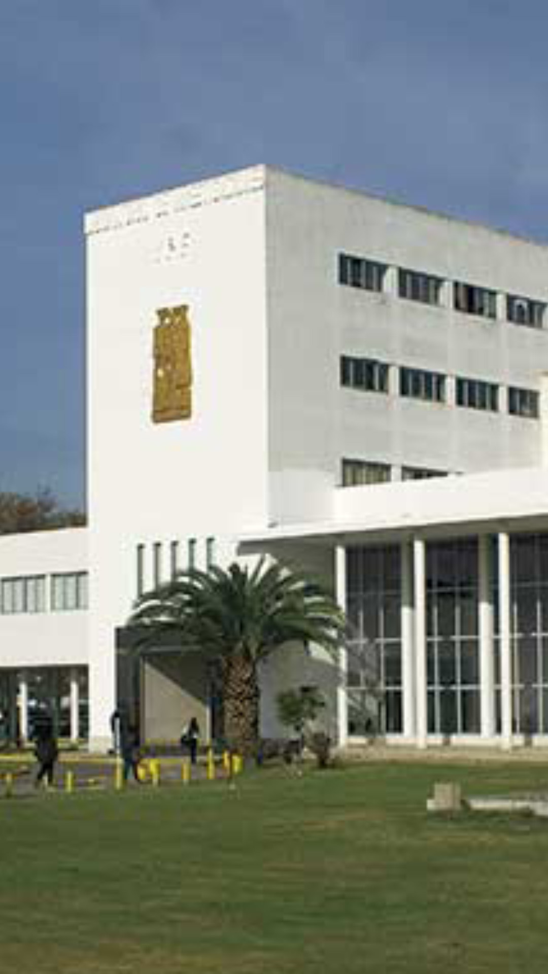 Portada sede central de la Universidad Nacional Arturo Jauretche. La foto es de día, exterior y es un plano general del edificio tomada desde lejos. El edificio es todo blanco con ventanales grandes y columnas. Se ven a lo lejos ingresando alumnos al establecimiento que muestra también un campo verde con algunas plantas.